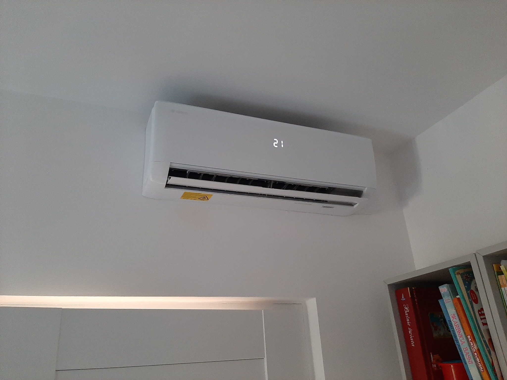Biały klimatyzator ścienny z wyświetlaczem temperatury (21 stopni Celsjusza), zamontowany w pokoju z białymi ścianami, widoczny fragment regału z książkami dla dzieci.
