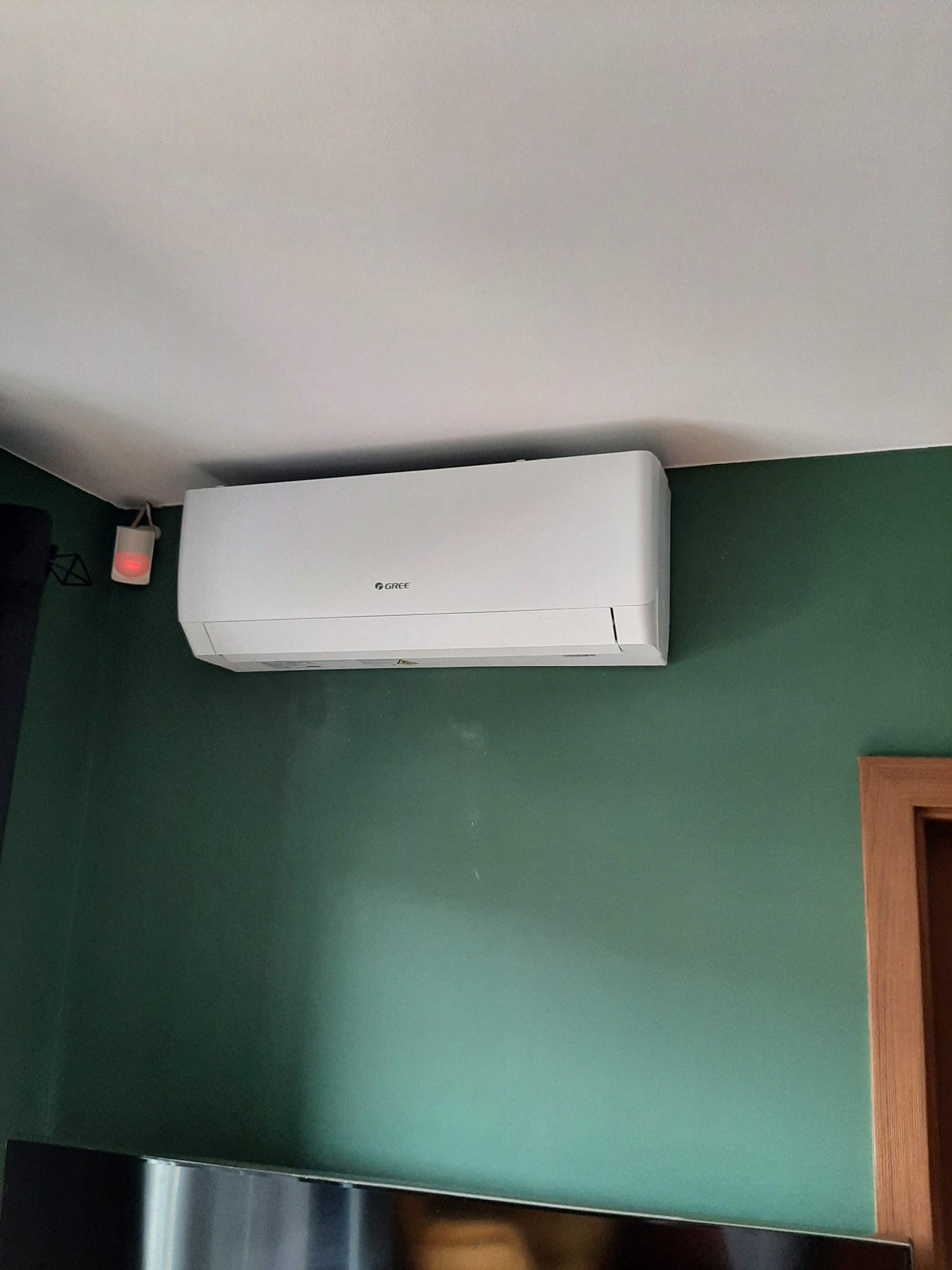 Biała jednostka klimatyzacyjna Gree zamontowana na zielonej ścianie w pomieszczeniu mieszkalnym, widoczny czujnik dymu na suficie.