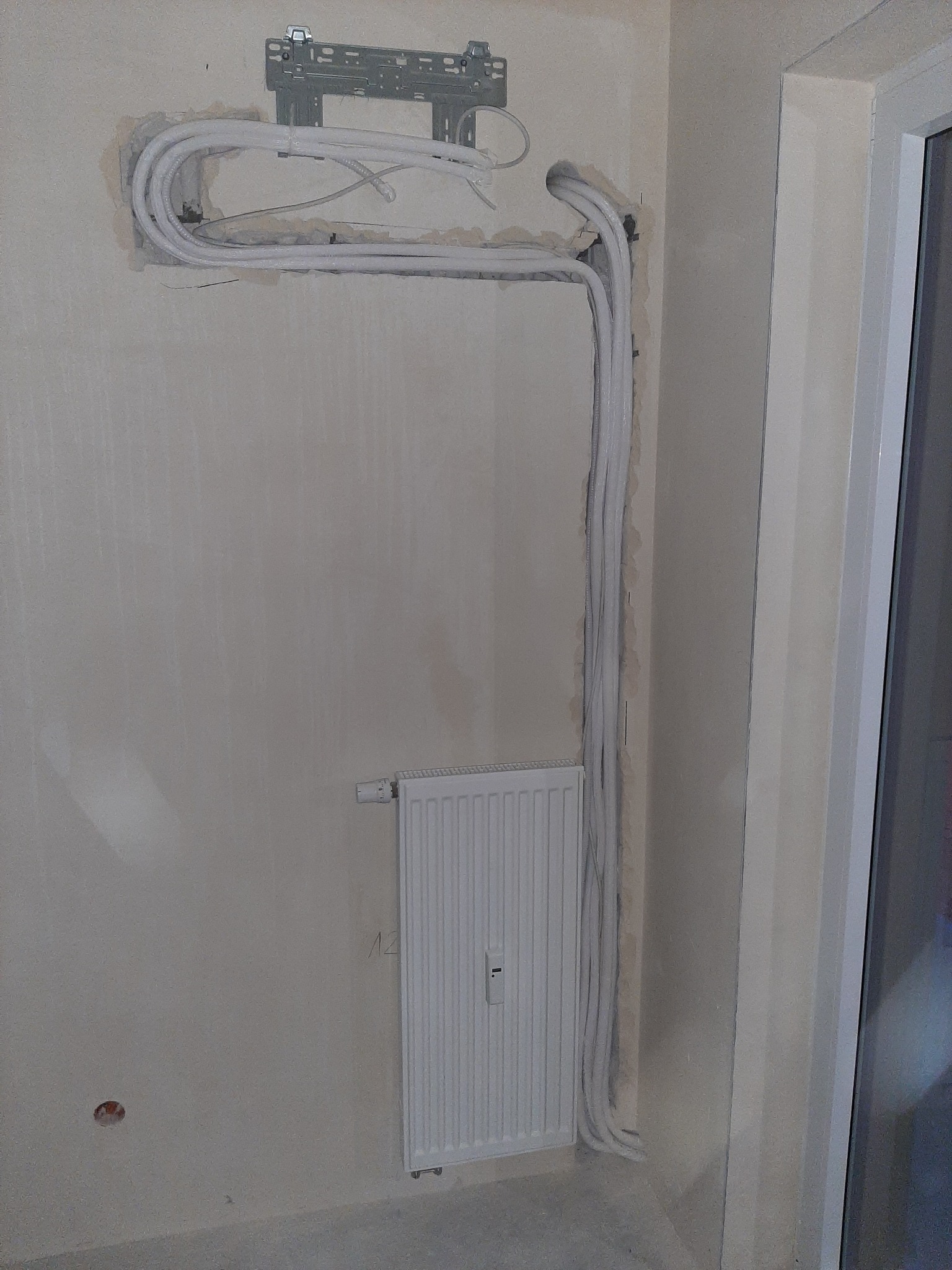 Wewnętrzna instalacja rur chłodniczych i elektrycznych pod klimatyzację, widoczny uchwyt montażowy, ściana w trakcie prac wykończeniowych, grzejnik panelowy.