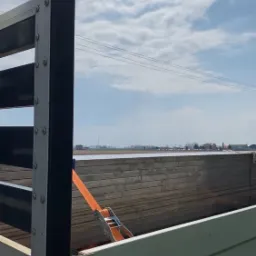 Ładunek płyt zabezpieczony pomarańczowym pasem w skrzyni ładunkowej samochodu na tle błękitnego nieba.