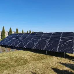 Panel fotowoltaiczny na metalowej konstrukcji, ustawiony na trawniku przed domem, w tle betonowy płot i kolumnowe tuje, błękitne niebo.
