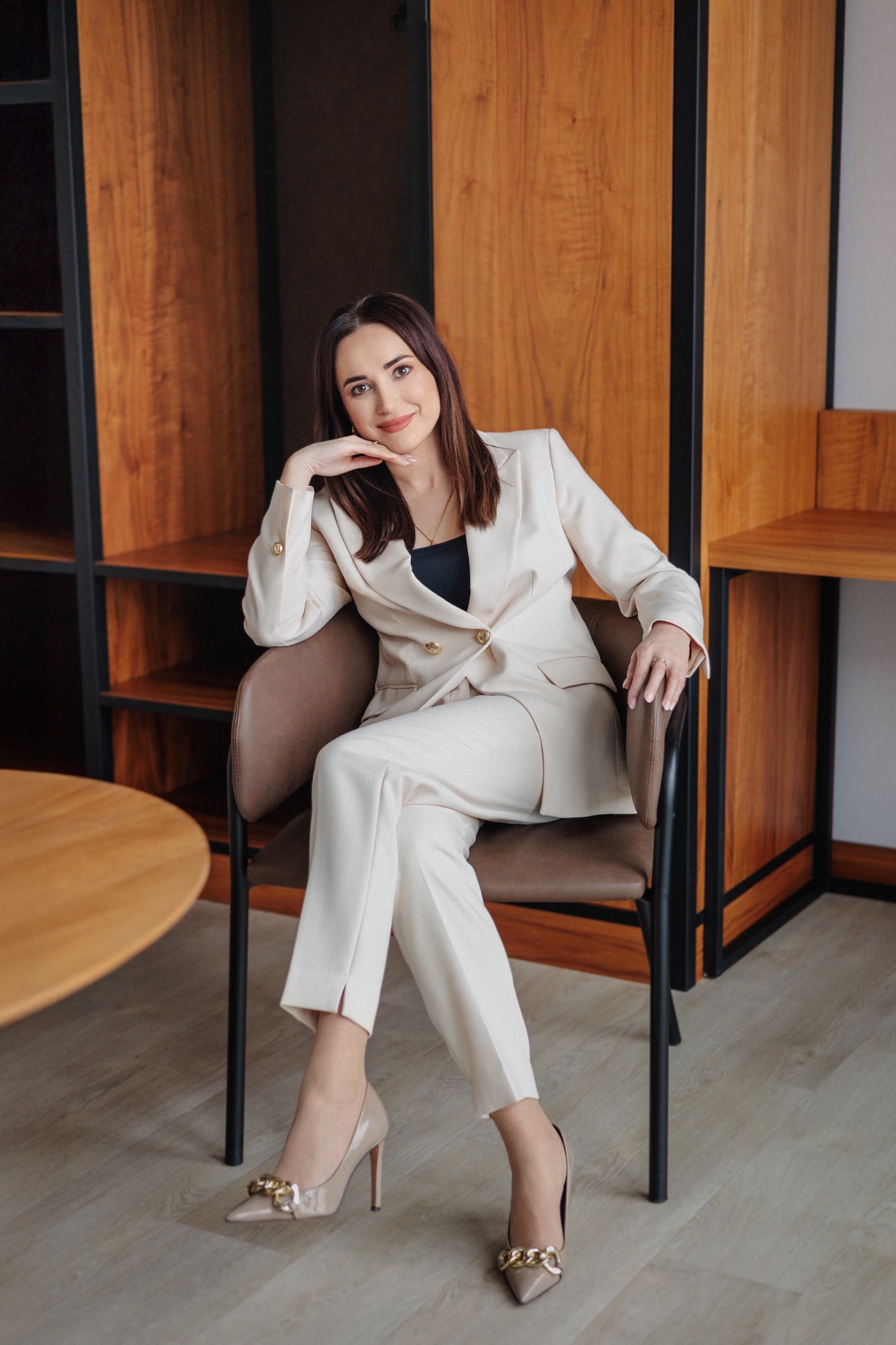 Portret kobiety w beżowym garniturze siedzącej na krześle w biurze. Uśmiechnięta, z ręką podpartą pod brodą, eleganckie szpilki. W tle regał z drewnianymi elementami.