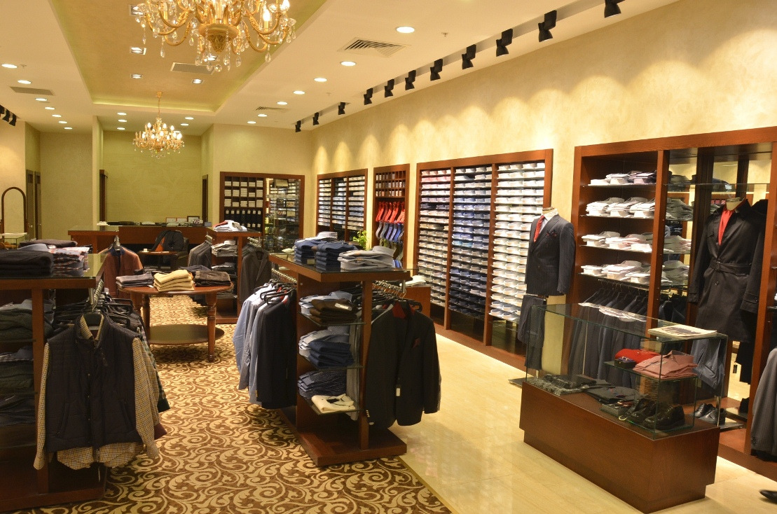 Wnętrze eleganckiego sklepu odzieżowego z klasycznymi ubraniami, oświetlonego kryształowymi żyrandolami, z ekspozycją koszul w szafach wnękowych i ubraniami na wieszakach na drewnianych regałach.