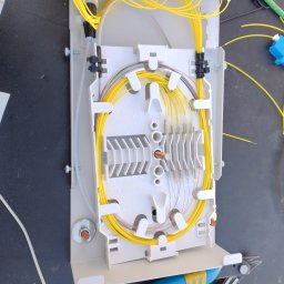 SignalPro - Rozdzielnica światłowodowa z żółtymi kablami wewnątrz, zamontowana na czarnej powierzchni. Widoczne narzędzia: szczypce z niebieskimi rączkami.