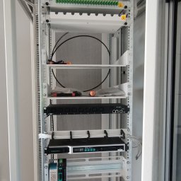 SignalPro - Wnętrze szafy rack z zamontowanymi urządzeniami sieciowymi: patch panel światłowodowy, router, zasilacz. Widoczne kable i narzędzia.