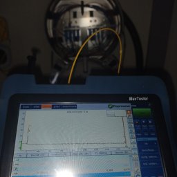 SignalPro - Tester światłowodowy EXFO z widocznym wykresem pomiarowym OTDR, powyżej studzienka telekomunikacyjna z włóknami światłowodowymi. Analiza sieci, pomiary.