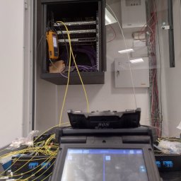 SignalPro - Montaż światłowodów w szafie rack. Widoczne patch panele, tester światłowodowy Fluke Networks i spawarka światłowodowa Fujikura 90S. Przewody światłowodowe w kolorze żółtym.