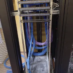 Szafa serwerowa z uporządkowanymi kablami sieciowymi w kolorach niebieskim i fioletowym, widoczne panele krosownicze i oznaczenia portów. Estetyczne ułożenie przewodów.