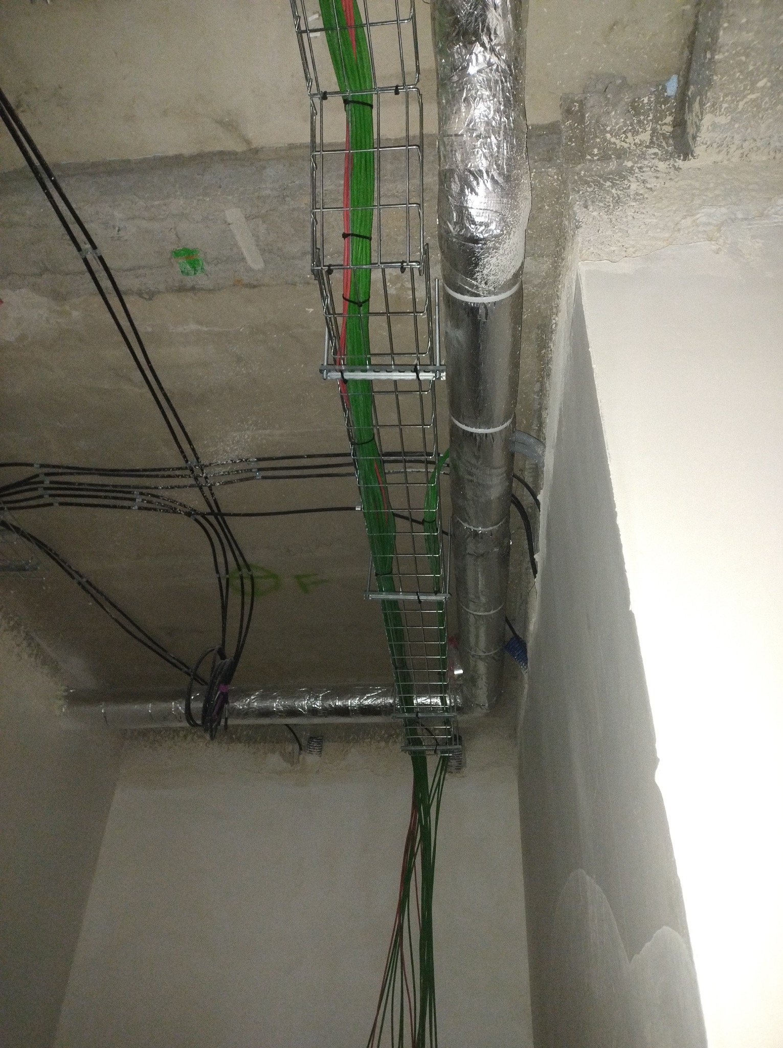 Kable w korytkach i rura wentylacyjna pod sufitem. Widoczne przewody w kolorze zielonym i czerwonym. Szare ściany i sufit.
