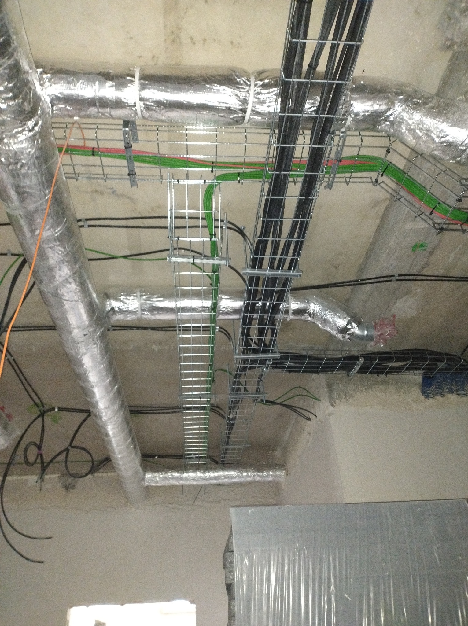 Instalacja kabli w korytach siatkowych pod sufitem, widoczne rury wentylacyjne w izolacji. Kable czarne, zielone i pomarańczowe. Surowy betonowy strop.