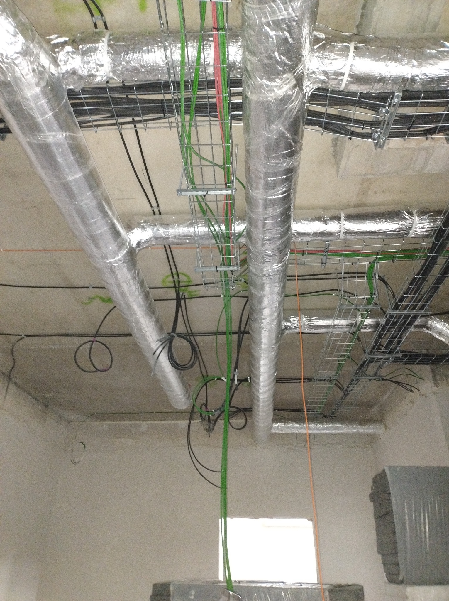 Widok na sufit z instalacją: rury w srebrnej izolacji, korytka kablowe z przewodami (zielone, czarne, pomarańczowe). Stan surowy, brak wykończenia ścian.