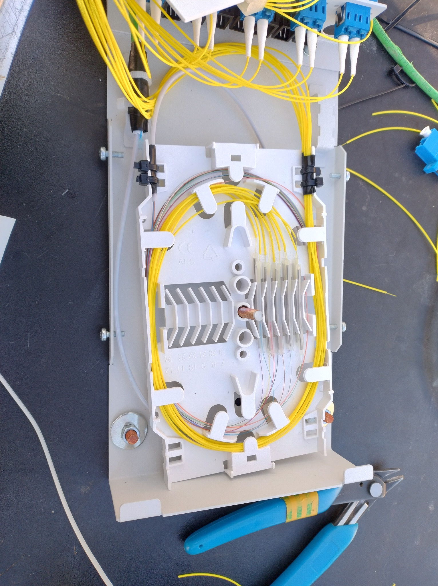 Rozdzielnica światłowodowa z żółtymi kablami wewnątrz, zamontowana na czarnej powierzchni. Widoczne narzędzia: szczypce z niebieskimi rączkami.