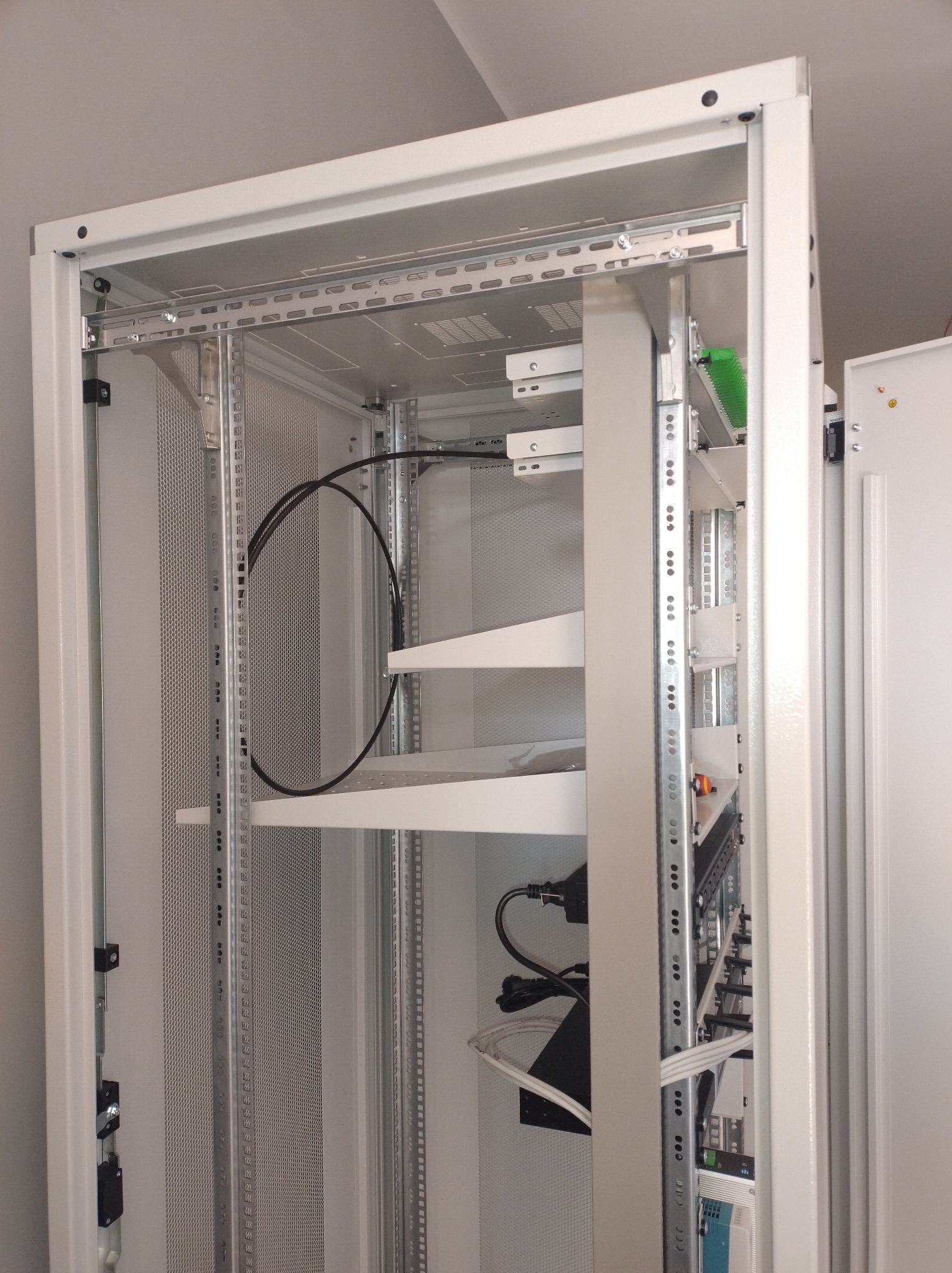 Wnętrze szafy serwerowej z półkami, okablowaniem i perforowanymi panelami bocznymi. Widoczne elementy montażowe i zasilające. Ujęcie z bliska, perspektywa od dołu.