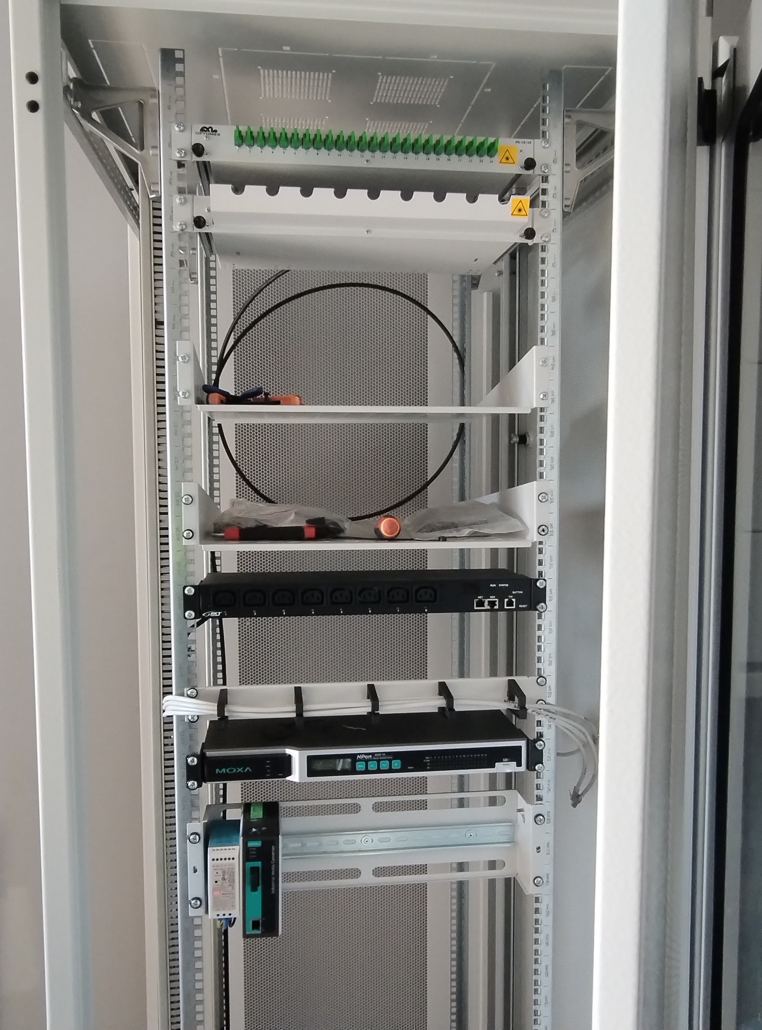 Wnętrze szafy rack z zamontowanymi urządzeniami sieciowymi: patch panel światłowodowy, router, zasilacz. Widoczne kable i narzędzia.