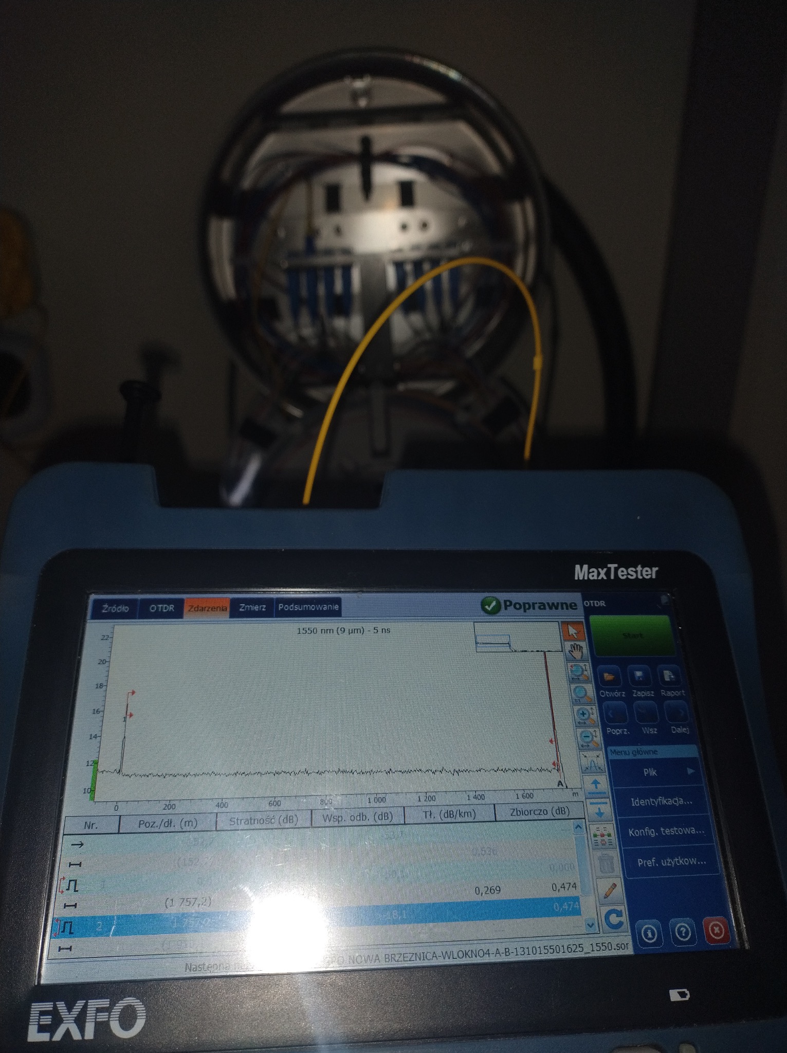 Tester światłowodowy EXFO z widocznym wykresem pomiarowym OTDR, powyżej studzienka telekomunikacyjna z włóknami światłowodowymi. Analiza sieci, pomiary.