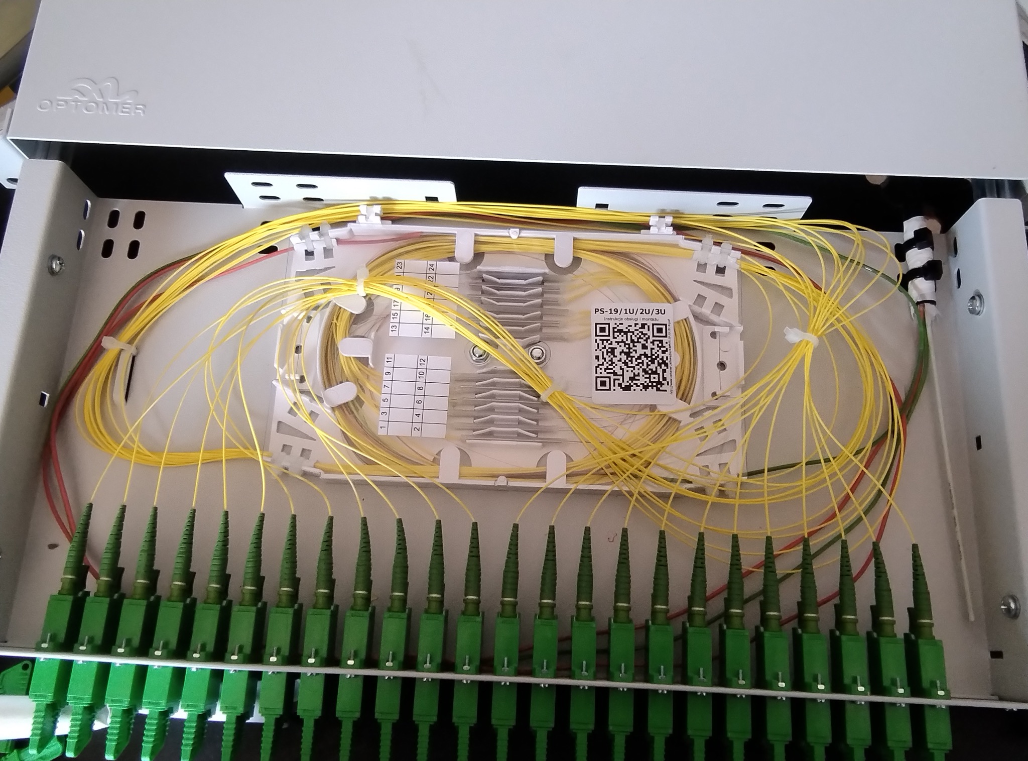 Rozdzielnica światłowodowa Optomer z precyzyjnie ułożonymi, żółtymi kablami. Widoczne zielone złącza i etykiety z numeracją. Estetyczne i uporządkowane okablowanie.