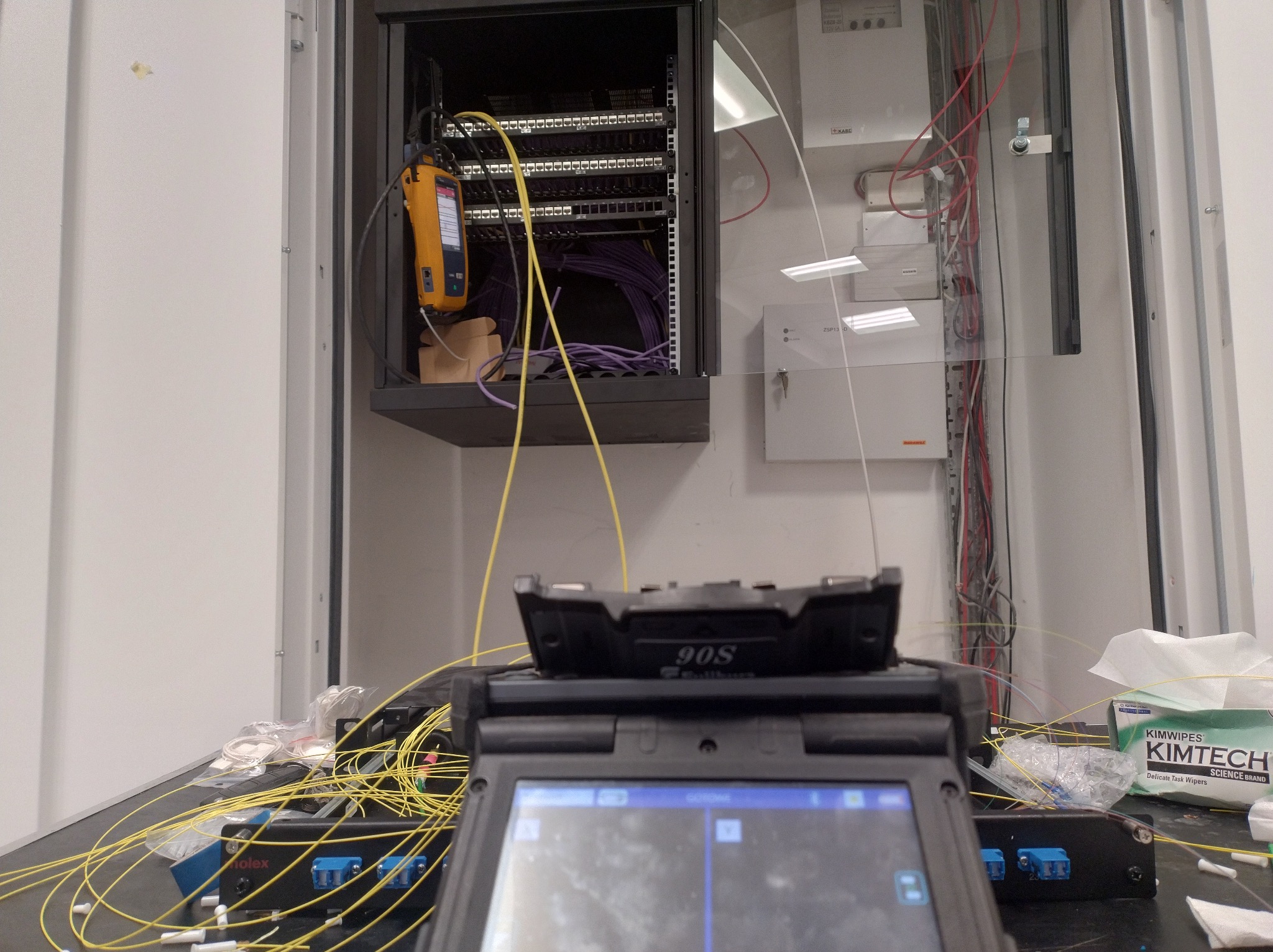 Montaż światłowodów w szafie rack. Widoczne patch panele, tester światłowodowy Fluke Networks i spawarka światłowodowa Fujikura 90S. Przewody światłowodowe w kolorze żółtym.