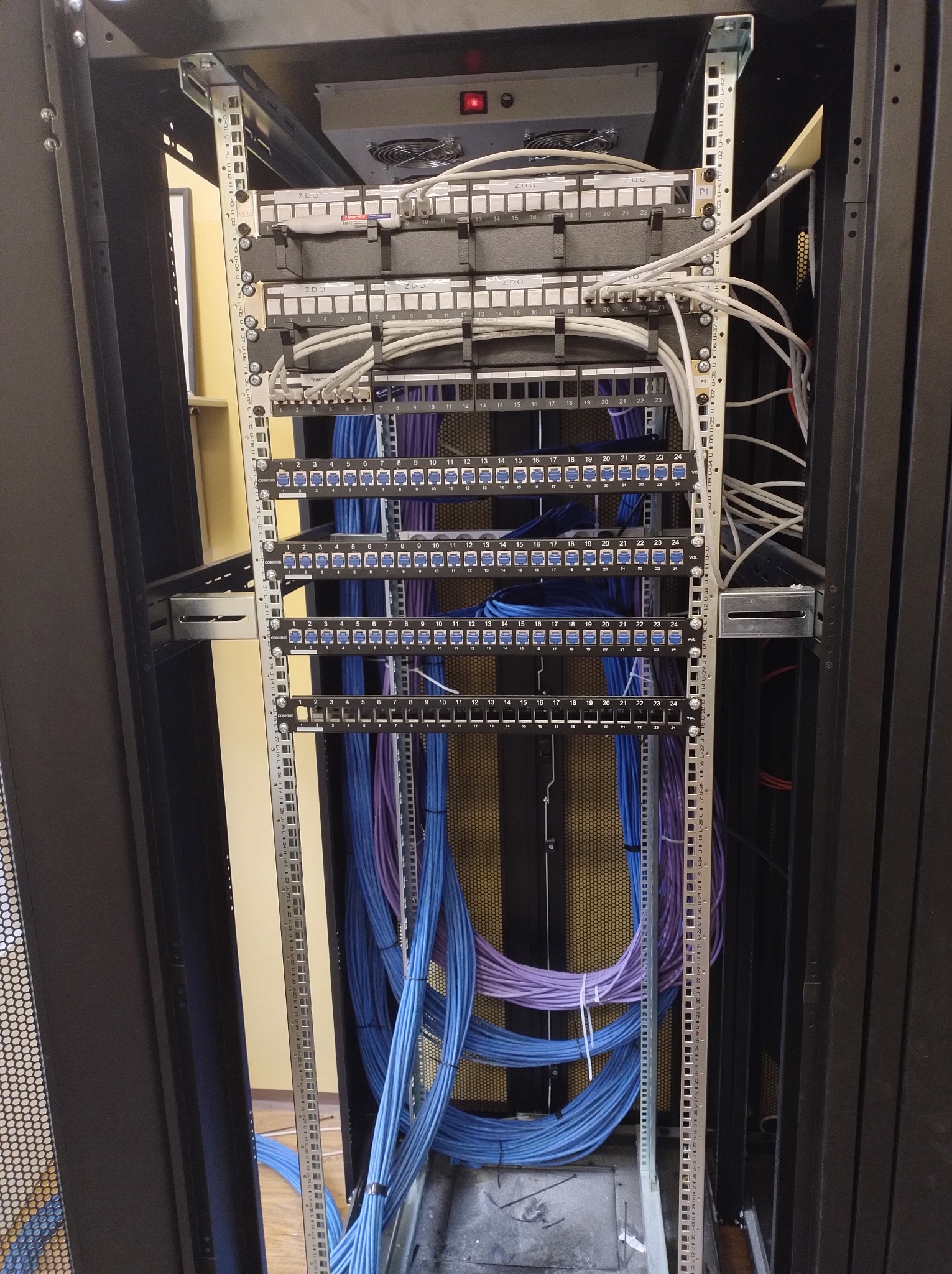 Szafa rack z panelami krosowymi i uporządkowanymi kablami patchcord, przewody w kolorze niebieskim i fioletowym, widoczne oznaczenia portów.