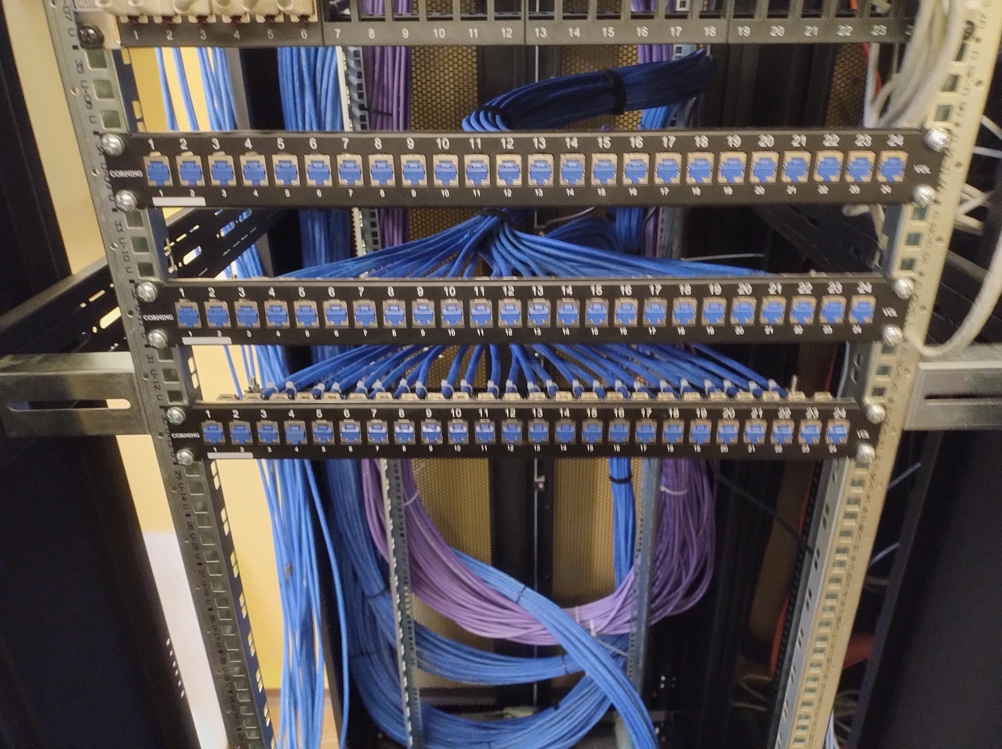 Panel krosowniczy z kablami sieciowymi w kolorach niebieskim i fioletowym, zainstalowany w szafie rack. Widoczne oznaczenia portów i metalowa konstrukcja montażowa.