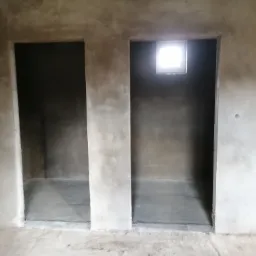 Surowe wnętrze z dwoma otworami drzwiowymi i oknem, ściany pokryte świeżym tynkiem, widoczne niedoskonałości i nierówności powierzchni.