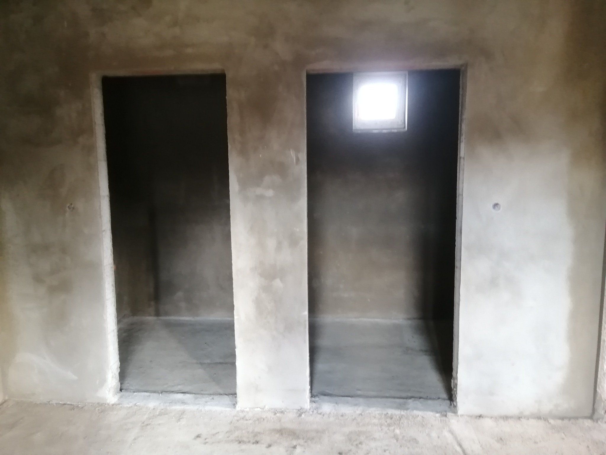 Surowe wnętrze z dwoma otworami drzwiowymi i oknem, ściany pokryte świeżym tynkiem, widoczne niedoskonałości i nierówności powierzchni.