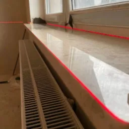Montaż parapetu okiennego z użyciem poziomicy laserowej, widoczny grzejnik i surowe wykończenie ściany.
