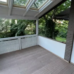 Zadaszony taras z drewnianą podłogą, białą balustradą i oknami dachowymi, widok na zieleń ogrodu.