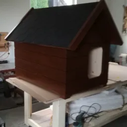 Brązowa buda dla psa z czarnym dachem z gontu bitumicznego, stojąca na drewnianym stole warsztatowym na kółkach w warsztacie, widok z boku.