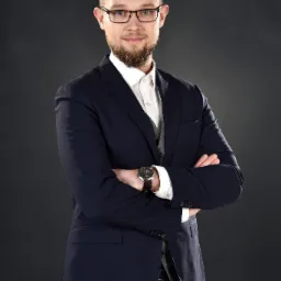 Portret młodego mężczyzny w okularach, granatowym garniturze i szarej kamizelce, z zegarkiem na skórzanym pasku, stojącego na ciemnoszarym tle z założonymi rękami.