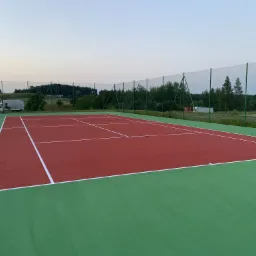 Widok na nowo wybudowany, czerwono-zielony kort tenisowy z siatką, otoczony zielenią i budynkami w oddali, sfotografowany o zmierzchu.