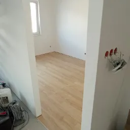 Wnętrze pokoju z drewnopodobną podłogą w trakcie wykańczania, widoczne niedokończone gniazdko elektryczne i narzędzia budowlane w rogu.