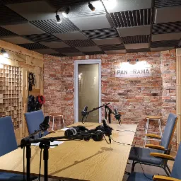 Wnętrze studia nagrań z mikrofonami, słuchawkami na stole, ścianą z czerwonej cegły z logo 'Panorama Studio Nagrań' i panelami akustycznymi na suficie.