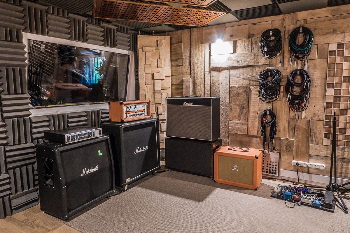 Wnętrze pomieszczenia z panelami akustycznymi i drewnianą ścianą, wzmacniacze Marshall i Orange, kable zwisające ze ściany, pedały gitarowe na podłodze.