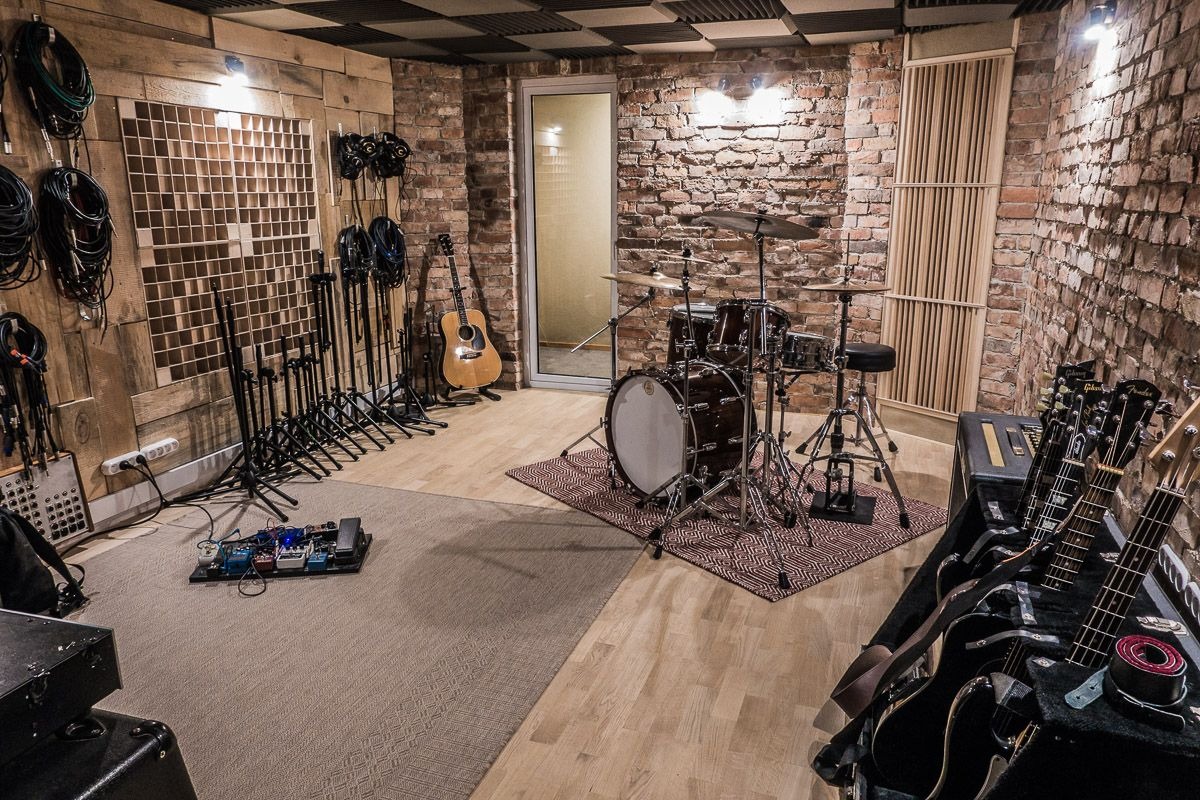 Wnętrze pomieszczenia z surowymi ceglanymi ścianami, panelami akustycznymi na suficie i ścianach, zestawem perkusyjnym na wzorzystym dywanie, gitarą akustyczną i elektrycznymi oraz efektami...