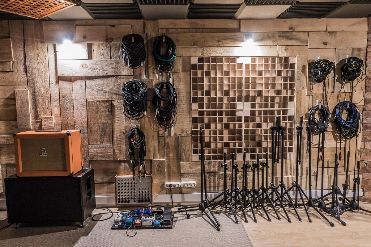 Wnętrze pomieszczenia z drewnianą ścianą, na której wiszą kable, słuchawki i znajduje się panel akustyczny z kwadratowymi przegródkami. Na podłodze wzmacniacz, kolumna głośnikowa, efekty gitarowe...