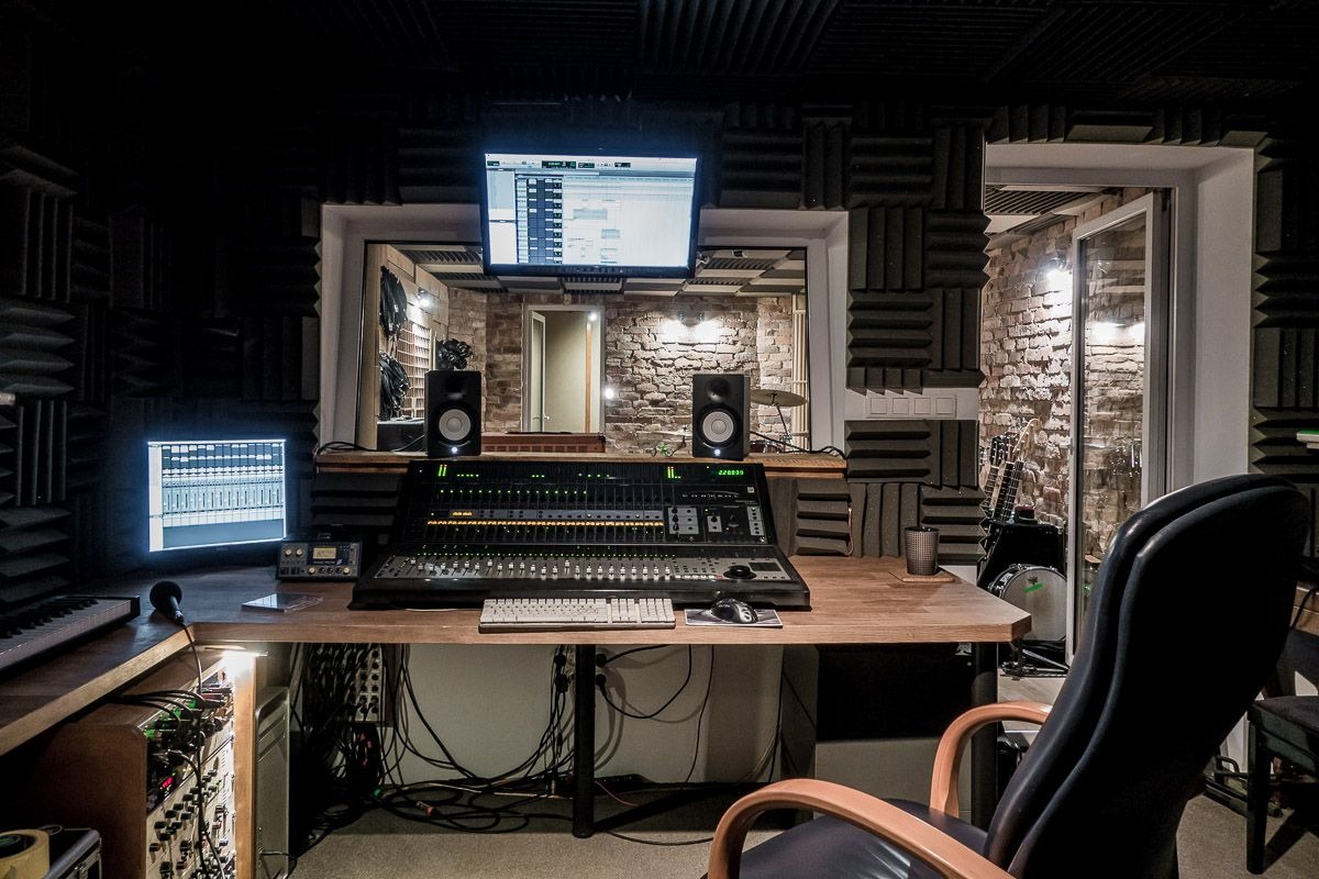 Profesjonalne pomieszczenie z wygłuszeniem akustycznym, mikserem dźwięku, monitorami odsłuchowymi, komputerem i instrumentami muzycznymi w tle za szybą.