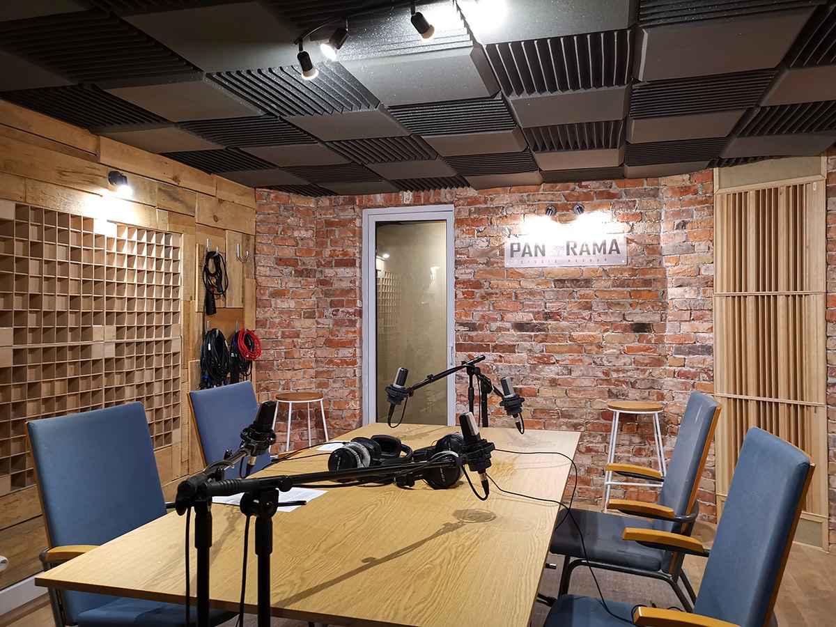 Wnętrze studia nagrań z mikrofonami, słuchawkami na stole, ścianą z czerwonej cegły z logo 'Panorama Studio Nagrań' i panelami akustycznymi na suficie.