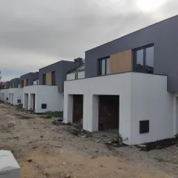 Rząd nowoczesnych domów szeregowych w trakcie wykończenia elewacji, z widocznym szarym tynkiem na górnej części i białym na dolnej, otwarte garaże z surową cegłą wewnątrz, teren wokół w trakcie...