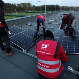 EN2 sp. z o.o. - Montaż paneli słonecznych na dachu budynku Legii Warszawa, widoczni pracownicy w czerwonych kamizelkach z logo firmy, w tle boisko sportowe.