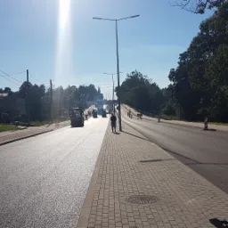 Świeżo położony asfalt na drodze w słoneczny dzień, z widocznymi walcami drogowymi i pracownikiem na chodniku z kostki brukowej, w tle drzewa i budynki.