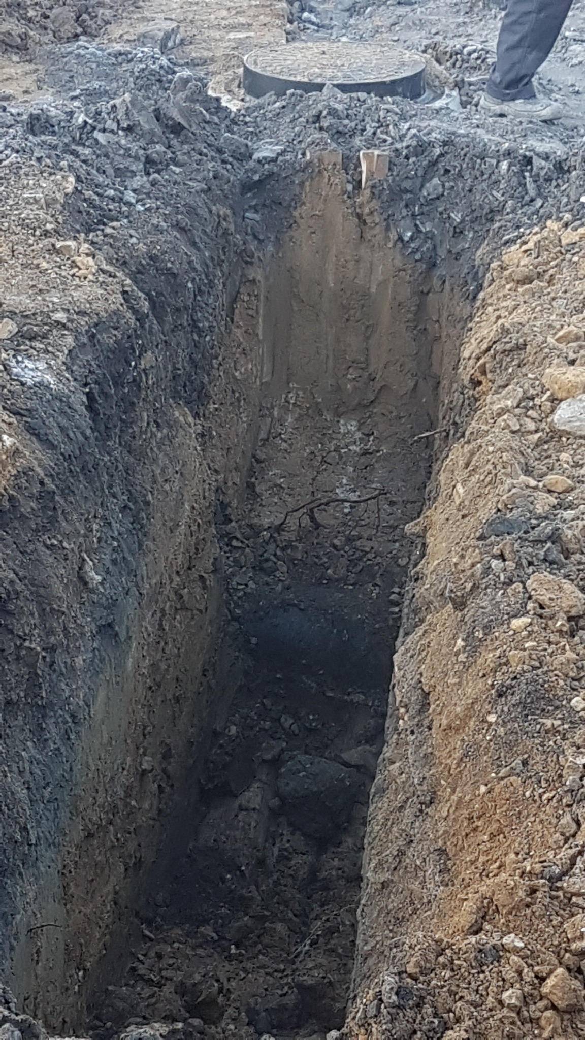 Głęboki wykop w ziemi, z widocznymi śladami po pracy koparki, obok właz kanalizacyjny i fragment buta osoby stojącej na górze.