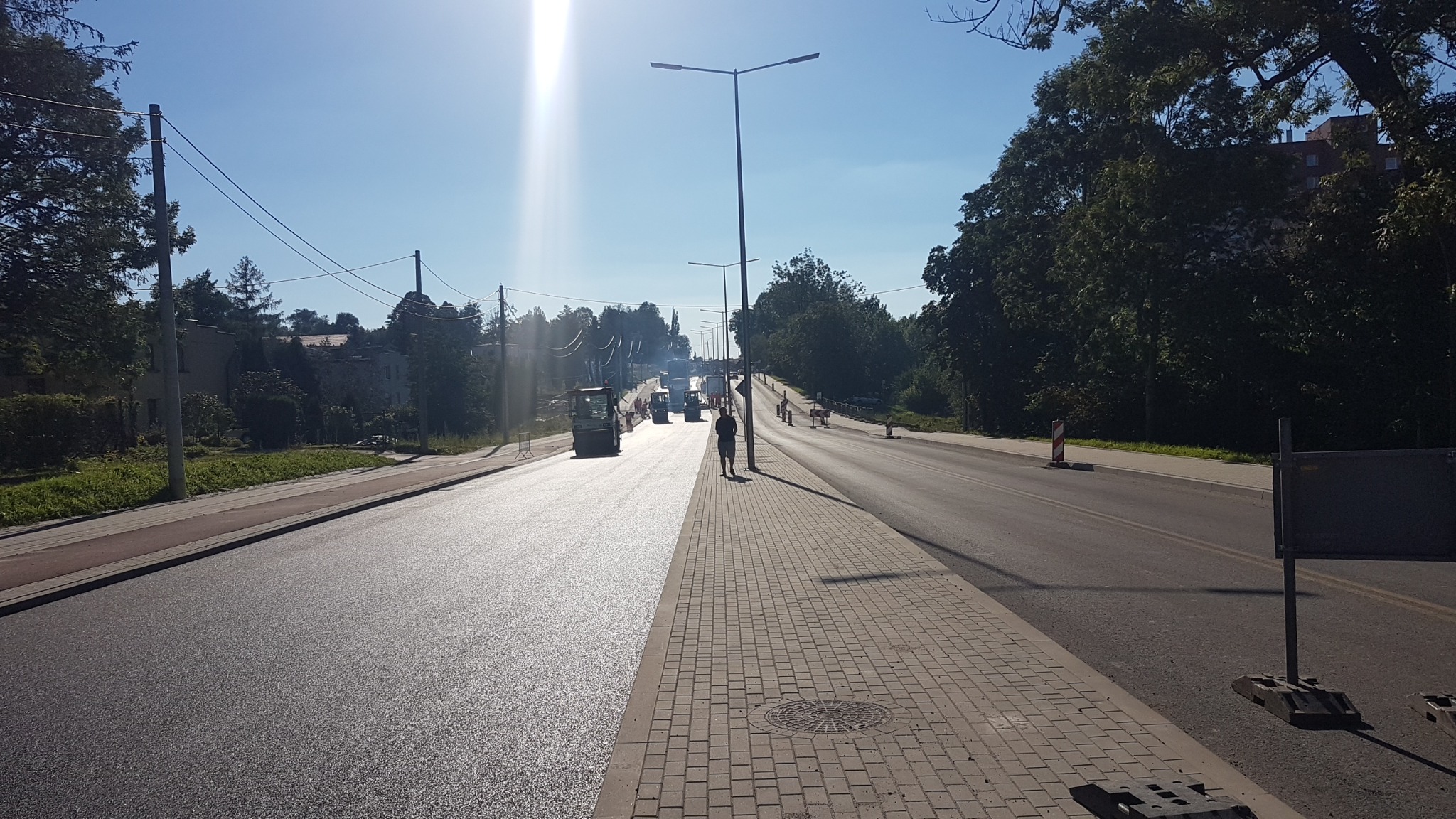 Świeżo położony asfalt na drodze w słoneczny dzień, z widocznymi walcami drogowymi i pracownikiem na chodniku z kostki brukowej, w tle drzewa i budynki.