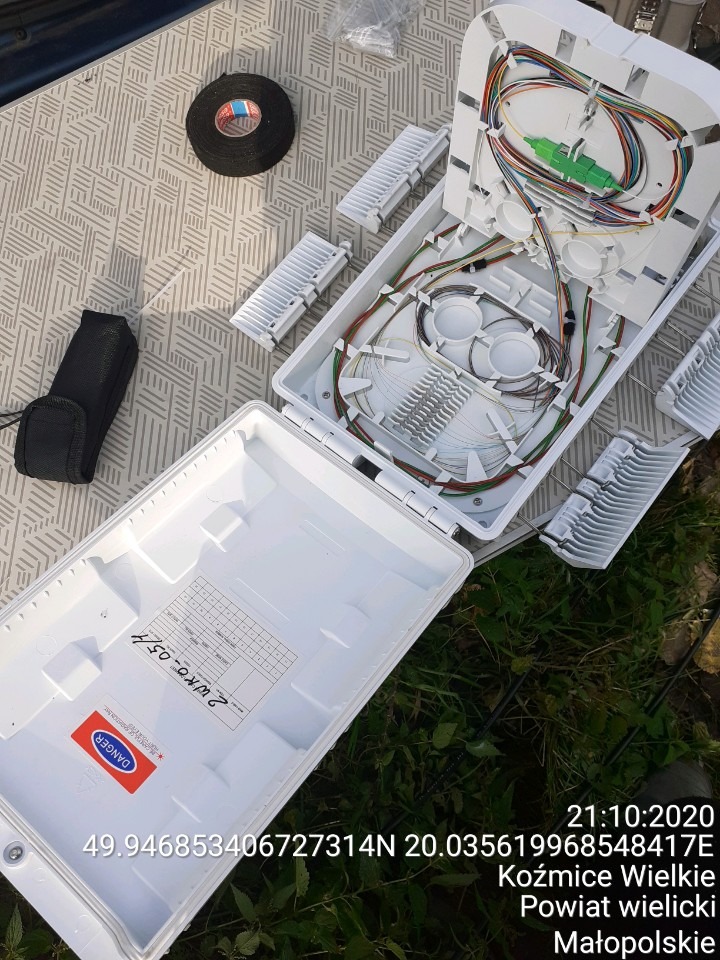 Otwarta biała skrzynka telekomunikacyjna z widocznymi światłowodami i elementami złącznymi na tle brązowej taśmy izolacyjnej i fragmentu trawy.