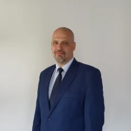 Portret mężczyzny w niebieskim garniturze i krawacie w kropki na jasnym tle.