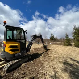 Żółto-czarna mini koparka Volvo ECR25D w trakcie prac ziemnych na wzgórzu pokrytym suchą trawą, błękitne niebo z obłokami w tle.
