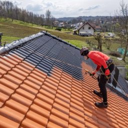 La Creative - Malowanie dachu z czerwonej dachówki na czarno przez pracownika w czerwonej koszuli, z użyciem specjalistycznego sprzętu natryskowego. Częściowo pomalowany dach na tle zieleni.