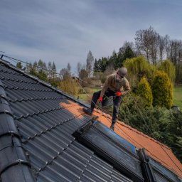 La Creative - Pracownik w uprzęży maluje dach z dachówki ceramicznej na czarno. Część dachu pomalowana, część w kolorze ceglastym. Widok z dołu na tle zieleni.