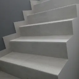 Wykończone schody wewnętrzne wyłożone jasnoszarymi płytkami ceramicznymi z delikatną fakturą, widok z dołu, szara ściana po lewej stronie.