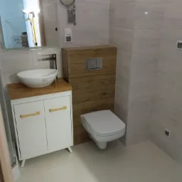 Nowoczesna łazienka z białą umywalką nablatową na szafce z drewnianym blatem, zabudowana toaleta z drewnianym frontem, lustro z oświetleniem widoczne w odbiciu, wykończenie w jasnych płytkach...