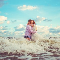 Janusz i Agata - Para młoda w objęciach w falach morskich; panna młoda w białej sukni, pan młody w mokrej, białej koszuli i czarnych spodenkach, niebo z chmurami w tle.